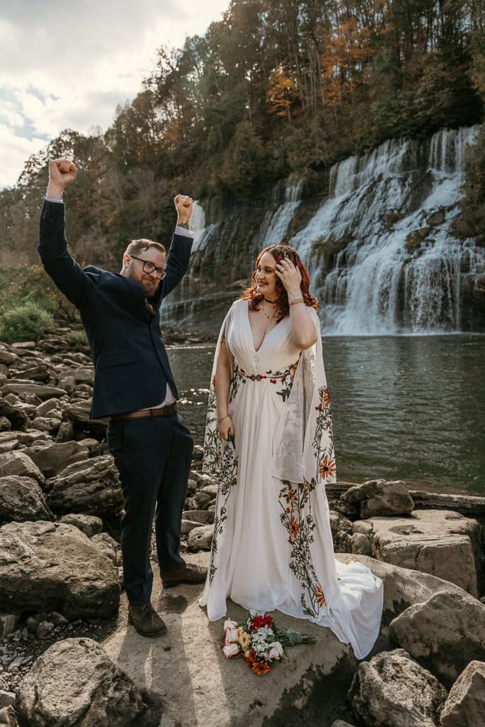 A man celebrates that he just got married by throwing his hands in the air. His wife laughs as she looks on. A waterfall is behind them.