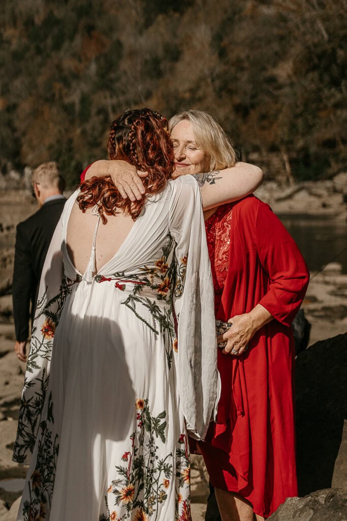A woman closes her eyes and embraces a bride with a smile on her face. They are outdoors.