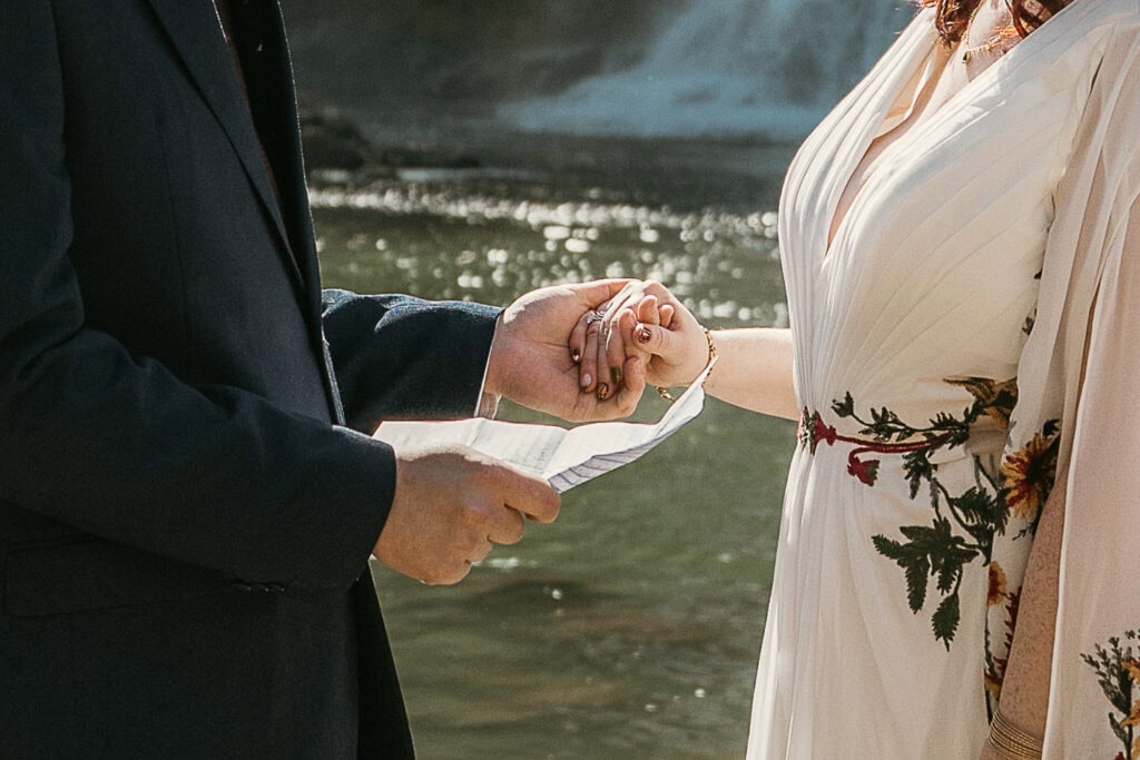 A man holds a woman's hand with one hand and in the other he holds a paper with his vows. Water is in the background.