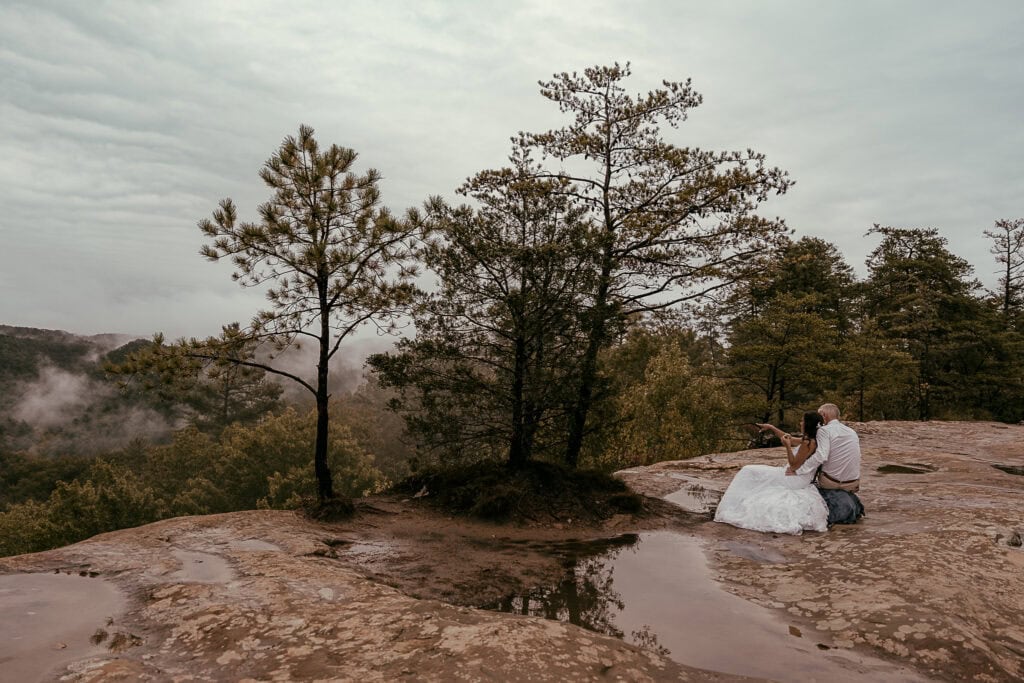 An eloping couple looks out in the distance from an overlook during their picnic.
