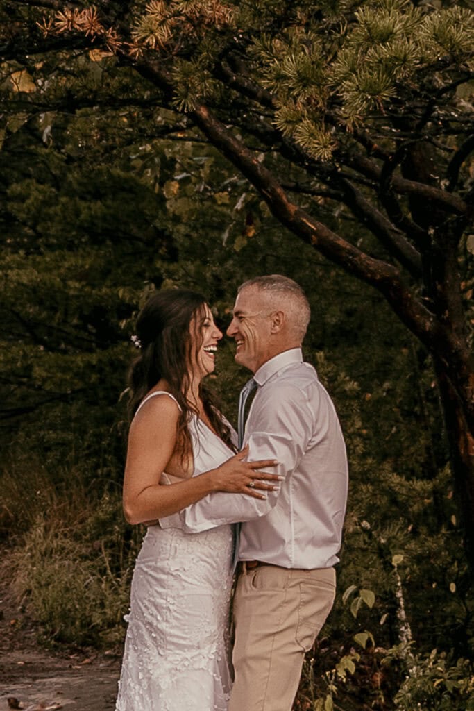 A couple holds each other while laughing. They are outdoors in wedding attire.