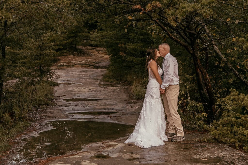 A newly married couple share a kiss. There is a puddle in the rocks and trees surround them.