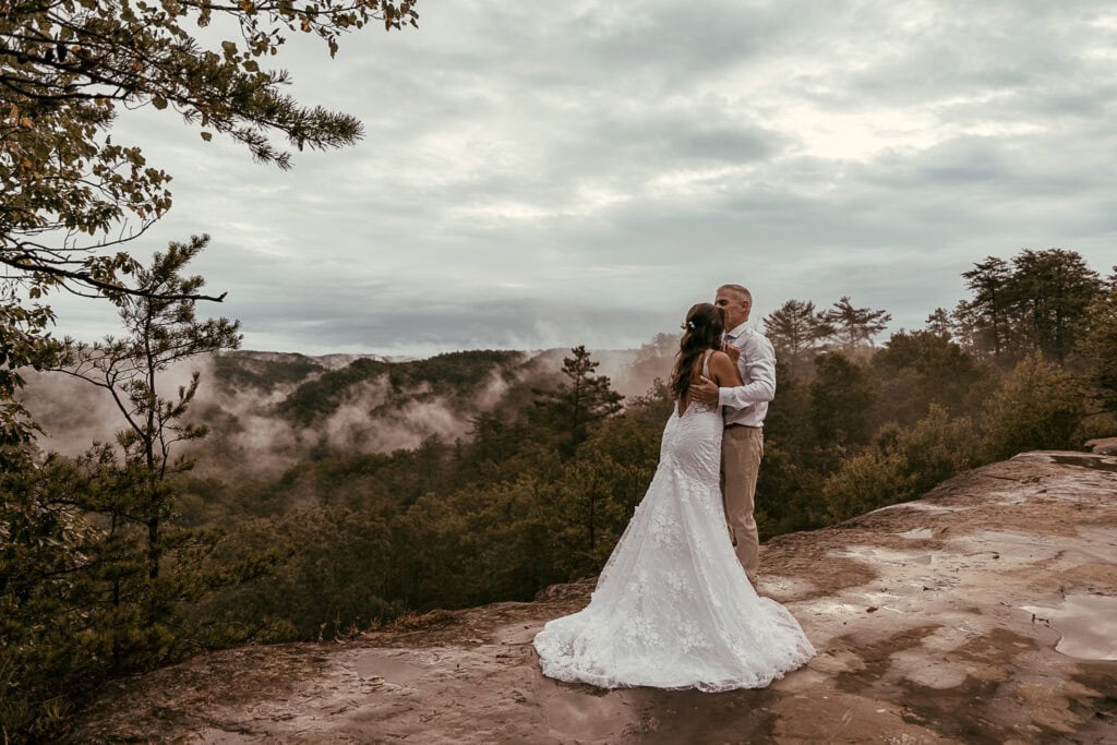 A couple in wedding attire stand on an overlook. A foggy valley is in the background.