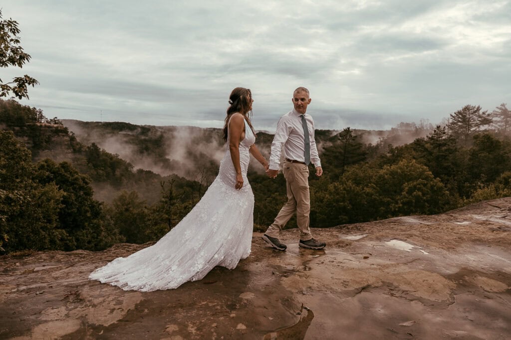 A couple walks hand in hand on a rock overlook during their elopement. A foggy valley is in the background.