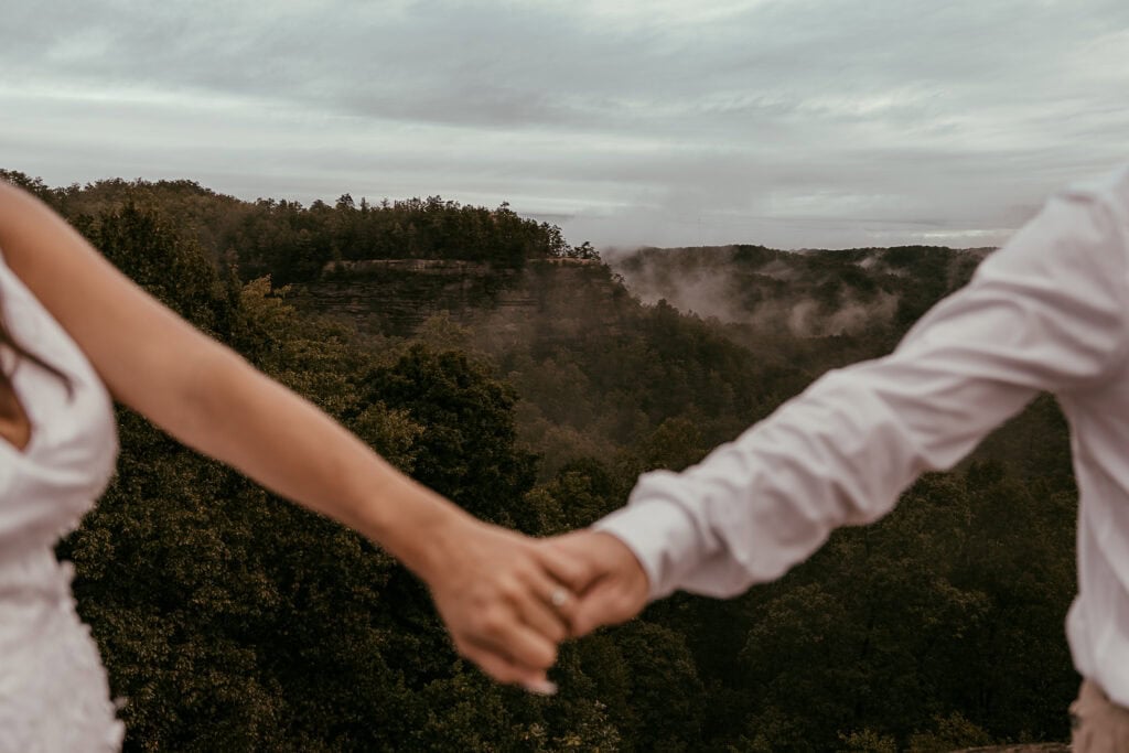 A closeup of a couple holding hands and rock formations and fog are behind them.