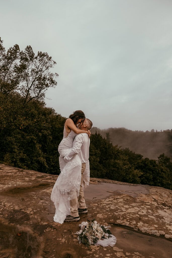 A groom picks his bride up for a kiss while they stand on top of Natural Bridge. It is foggy.