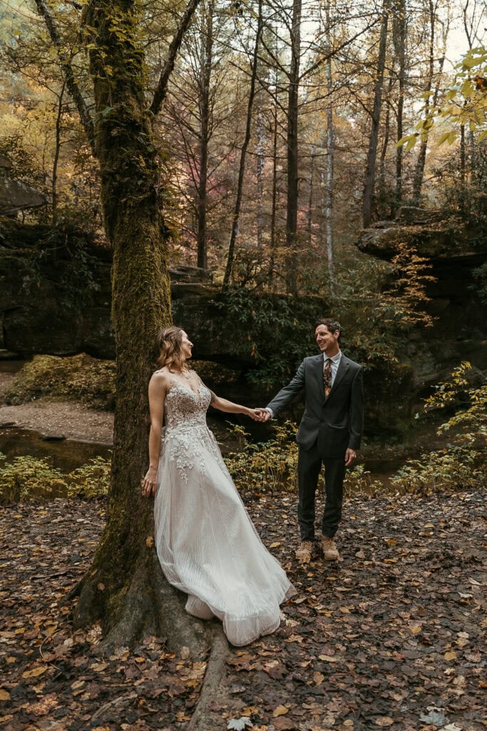 A bride leans against a tree while holding the groom's hand. It's fall and they're in the forest.